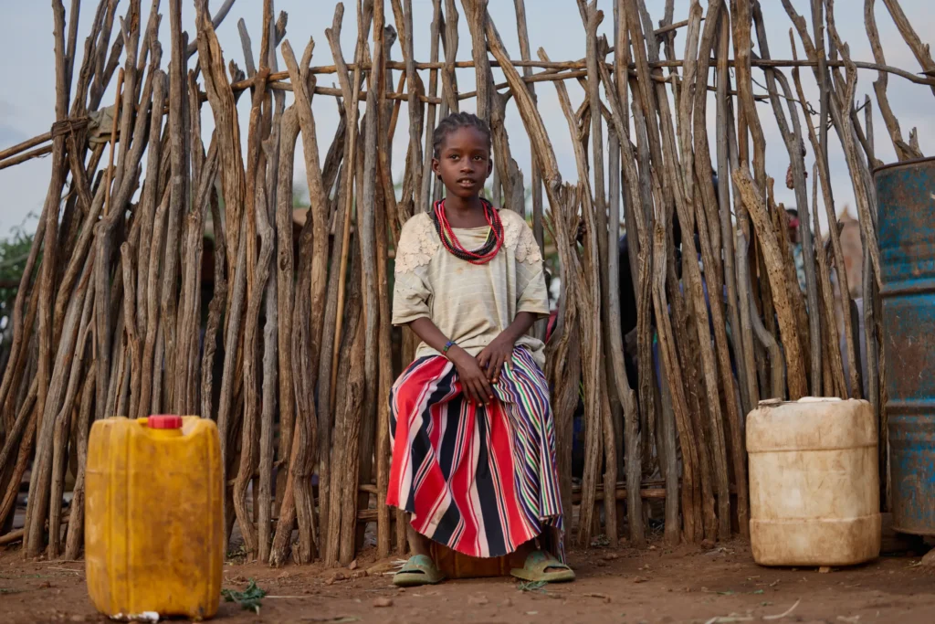 Hamar girl sitting near water containers in front of wooden fence in Ethiopia | Michaela Pupíková Photography