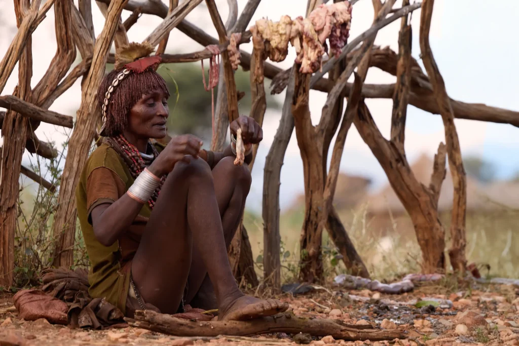 Hamar woman sitting beneath wooden rack with hanging meat in village, Ethiopia | Michaela Pupíková Photography