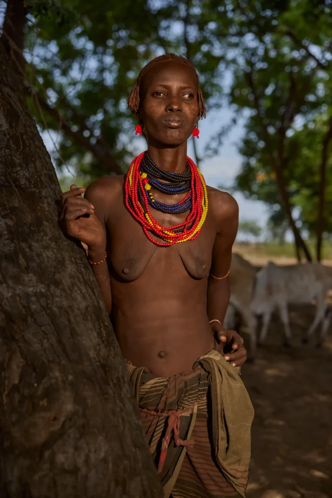 Dassanech woman wearing layered bead necklaces leaning against tree in Ethiopia | Michaela Pupíková Photography