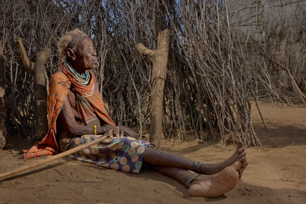 Old Nyangatom woman resting under stick fence with walking cane in traditional village, Ethiopia | Michaela Pupíková Photography